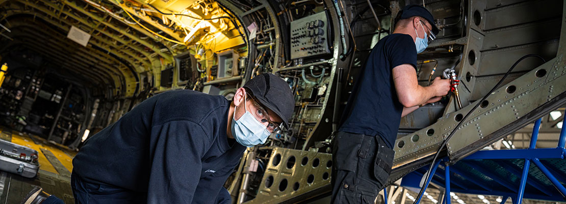 Two workers with bump caps, safety glasses, and masks working inside a fuselage